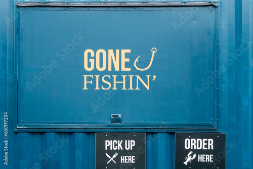 A blue metal wall window with gone fishing painted in yellow text and a fishing hook symbol.  Below the door there are two black signs with pick up here and order here. There's a knife and fork symbol