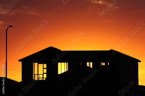 The silhouette of a two-story home under construction with the bright orange-yellow glow of the sunset as a backdrop photo