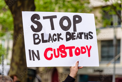 A homemade protest sign with the text 'Stop Black Deaths in Custody' is held up by a female hand