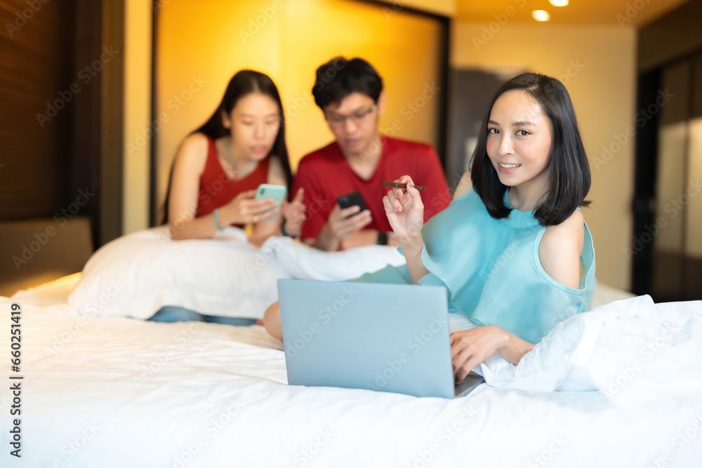 Asian business woman boss Lie down and look at work on a laptop, holding a pen. Behind him are co-workers working with smartphones. Wear a red shirt and a light green dress. In the hotel bedroom Asian business woman boss Lie down and look at work on a laptop, holding a pen. Behind him are co-workers working with smartphones. Wear a red shirt and a light green dress. In the hotel bedroom
