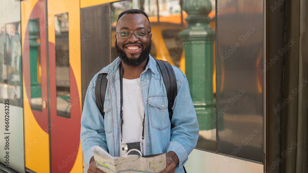 Happy Smiling young african american man in glasses with map at european terminal train station looking directly an the camera. - obrazy, fototapety, plakaty 