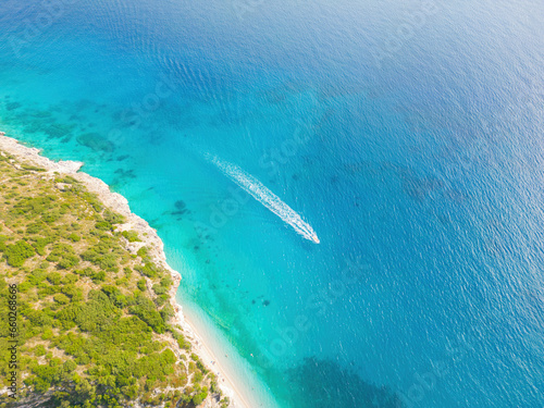 Aerial view of a clear blue sea, a boat leaving a trail, and a sunlit shoreline bordered by green vegetation. © ottaviocamb