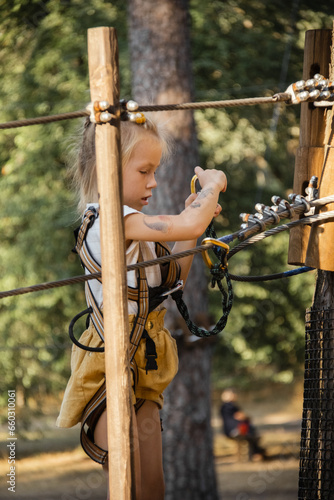 Cute little girl climbs and walks on a wooden path and platforms holding with a high ropes on tall green coniferous trees in rope park. Safety measures while having fun in an adventure rope park