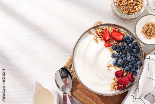 Bowl with yogurt and berries on board, jar and towel on light background, space for text