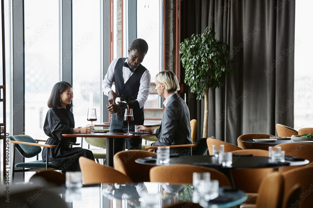 Interior of luxury restaurant with couple enjoying dinner date and server pouring red wine into glasses, copy space Interior of luxury restaurant with couple enjoying dinner date and server pouring red wine into glasses, copy space