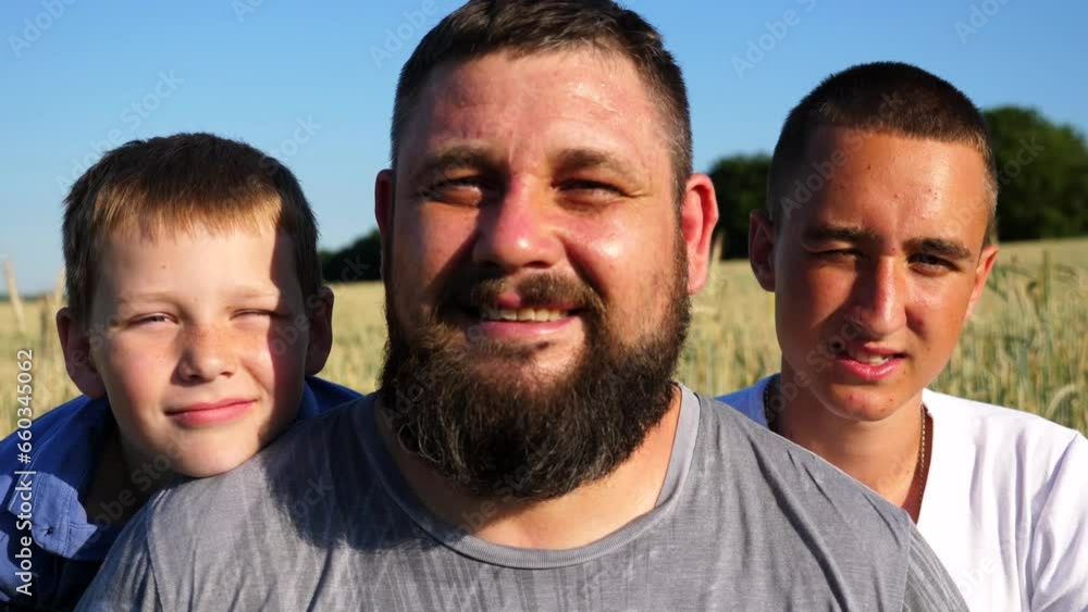 Young father with his two sons looks into camera against background of green field. Portrait of happy family at cultivated meadow. Close up emotions of dad and kids with positive expression on faces Young father with his two sons looks into camera against background of green field. Portrait of happy family at cultivated meadow. Close up emotions of dad and kids with positive expression on faces