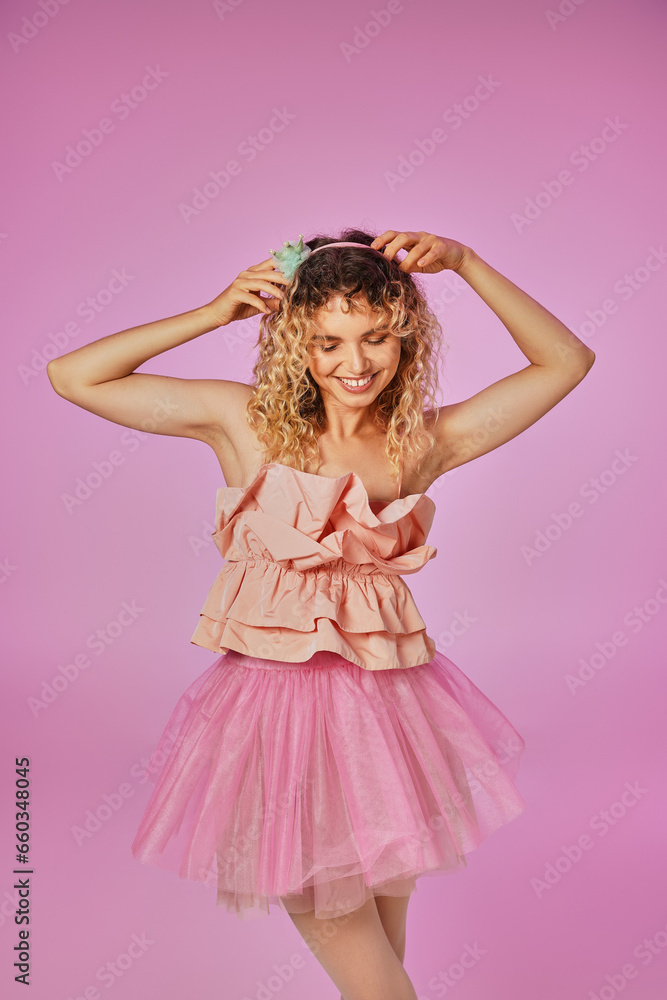 lovely curly haired tooth fairy in pink costume touching her headband and smiling on pink backdrop lovely curly haired tooth fairy in pink costume touching her headband and smiling on pink backdrop
