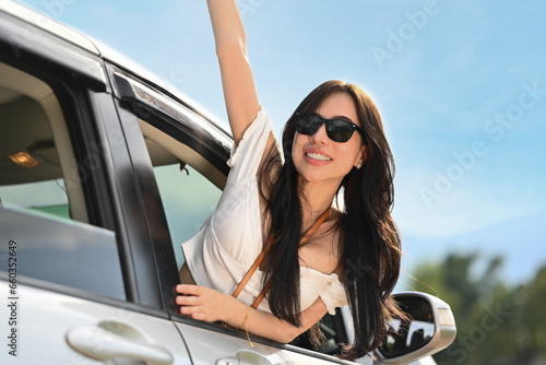 Carefree young woman in sunglasses leaning out of a car window, enjoying nature during summer spring road trip