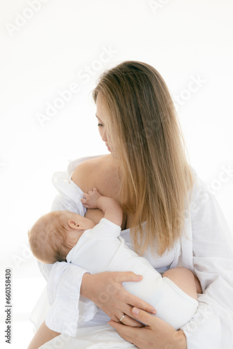 Young woman with baby in her arms. Blonde girl in white clothes smiling to your child on white background. Happy motherhood and breastfeeding concept. Photos with sun glare, soft focus, overexposure