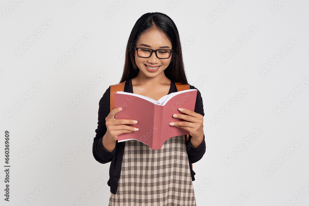 Smiling young Asian woman student in a casual shirt glasses and backpack standing while reading a study book isolated on white background. Education in high school university college concept Smiling young Asian woman student in a casual shirt glasses and backpack standing while reading a study book isolated on white background. Education in high school university college concept