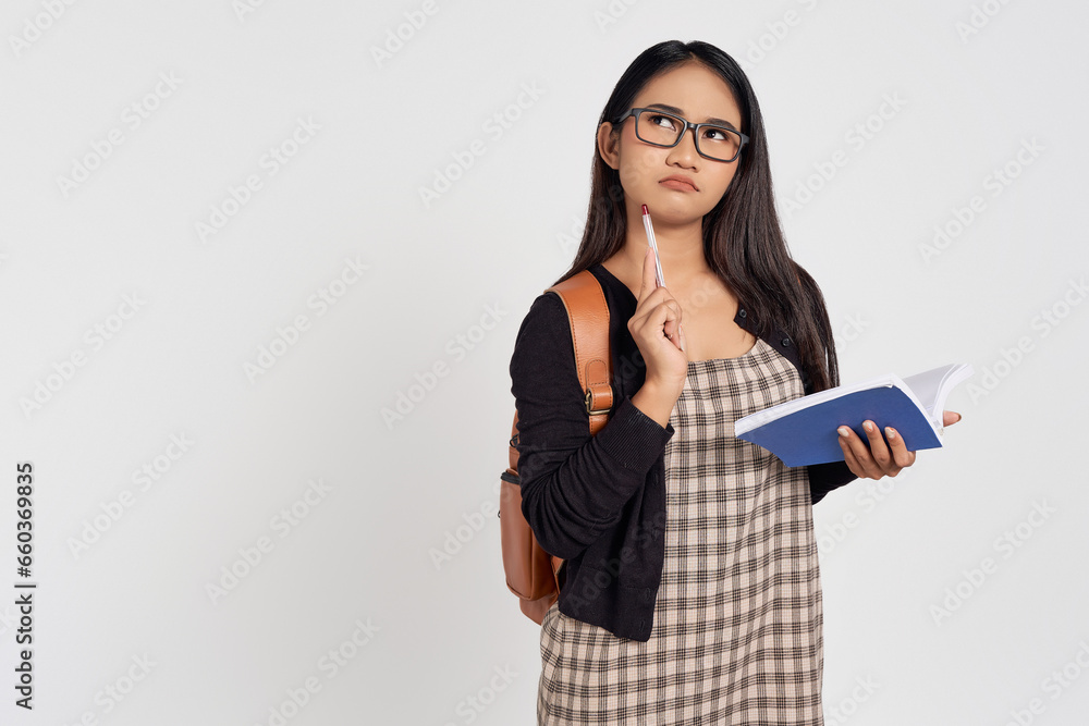 Pensive young Asian woman student in casual shirt glasses and backpack holding book and pen, thinking about something isolated on white background. Education in high school university college concept Pensive young Asian woman student in casual shirt glasses and backpack holding book and pen, thinking about something isolated on white background. Education in high school university college concept