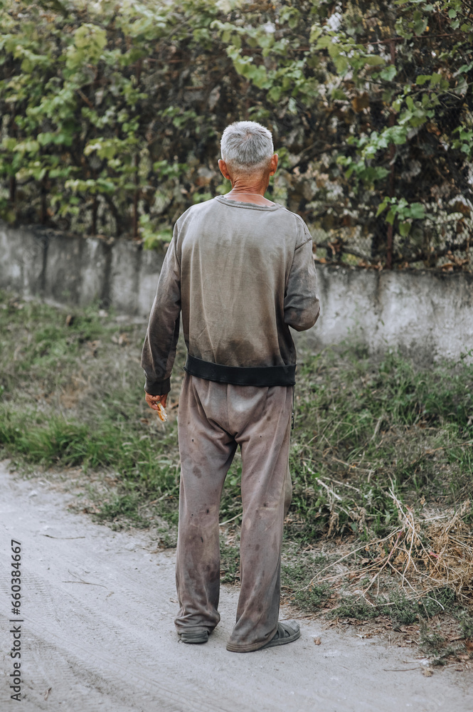 An old adult gray-haired thin homeless hungry man in dirty clothes walks along the street road in search of food. Photography, portrait.