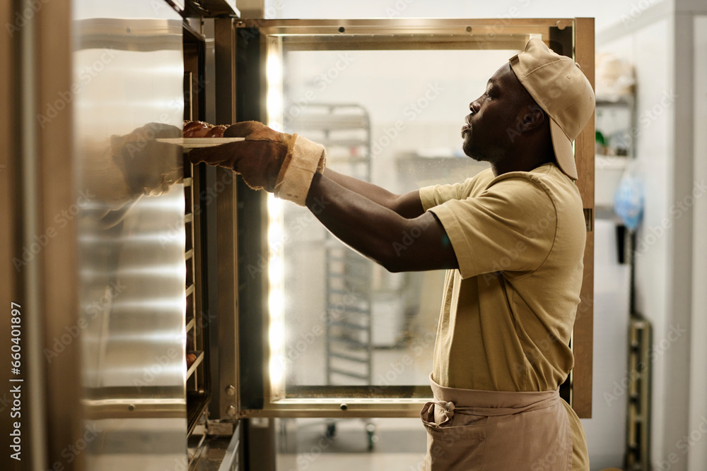 Side view portrait of Black young man putting tray with fresh breads in oven at bakery kitchen, copy space