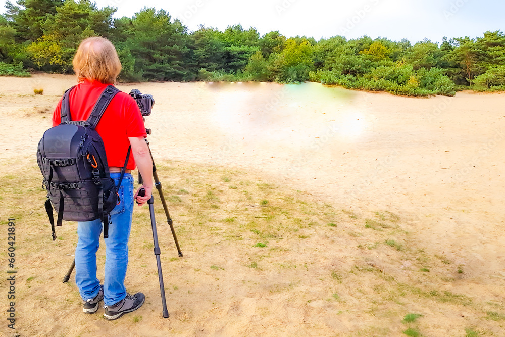 Rear view of a senior adult male photographer standing next to his camera on a tripod, dune landscape with green trees in blurred background, sunny spring day in Weert, North Limburg, Netherlands