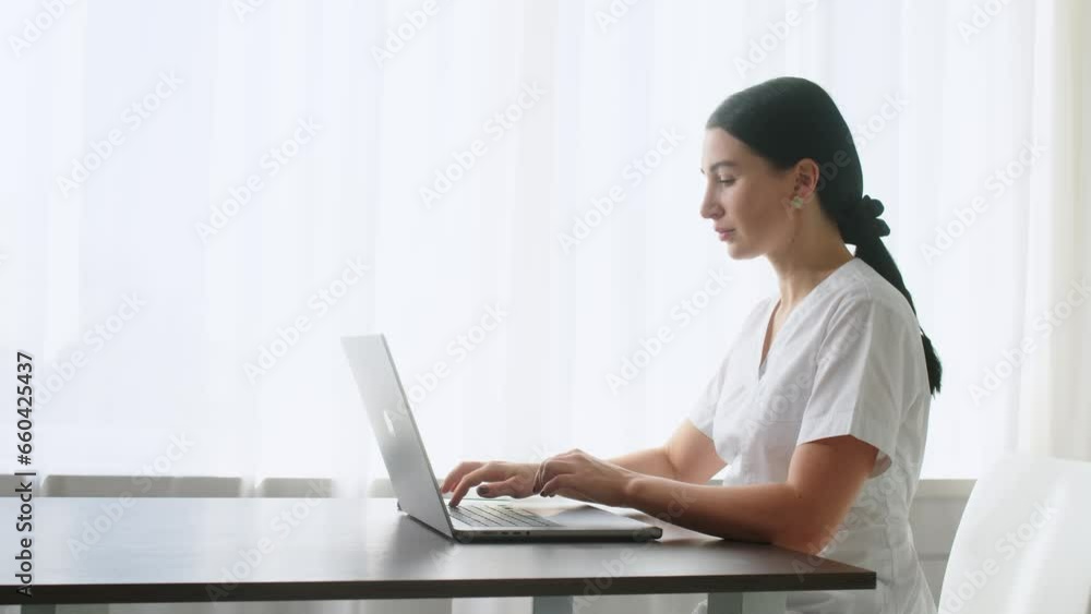 Close-up of a female family doctor working on a laptop computer in a clinic. A doctor in a white lab coat reviews a medical history at a table in a hospital office. Close-up of a female family doctor working on a laptop computer in a clinic. A doctor in a white lab coat reviews a medical history at a table in a hospital office.