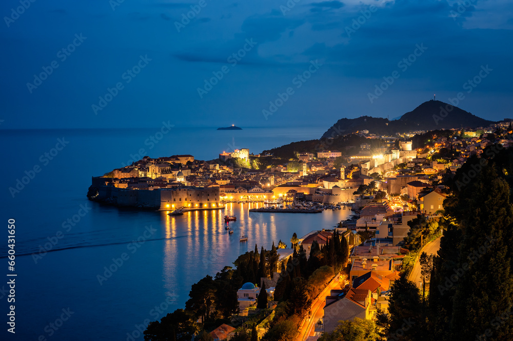 Amazing aerial panoramic view of the picturesque town of Dubrovnik with the old town, illuminated streets and buildings and marina with boats at night.
