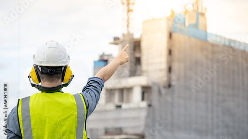 Asian male construction worker or site engineer man with green reflective vest and yellow safety helmet pointing at unfinished building structure and tower crane photo