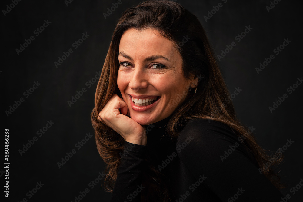 Portrait of an attractive middle aged woman with toothy smile wearing turtleneck sweater and sitting at isolated dark background. Portrait of an attractive middle aged woman with toothy smile wearing turtleneck sweater and sitting at isolated dark background.