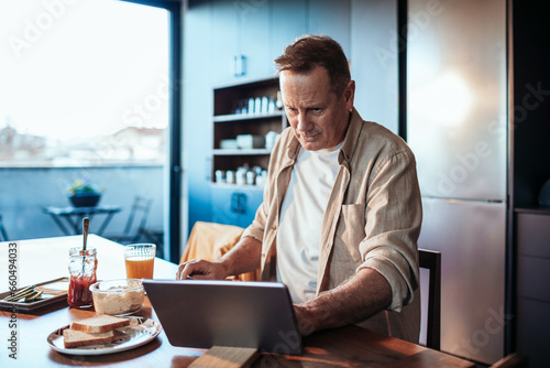 Middle aged man using the tablet while having breakfast in the kitchen photo