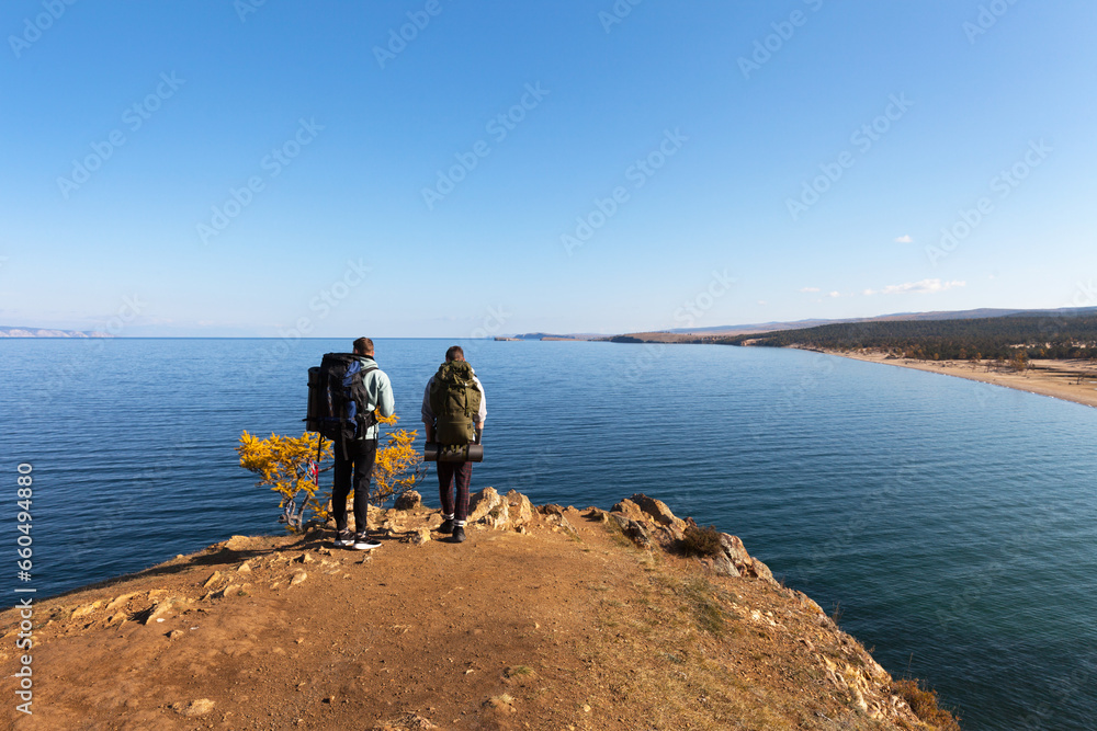 Baikal Lake in Autumn. Tourists with backpacks travel around Olkhon Island on foot and admire the beautiful view of Saraisky Bay from the cliff. Autumn active outdoors and recreation