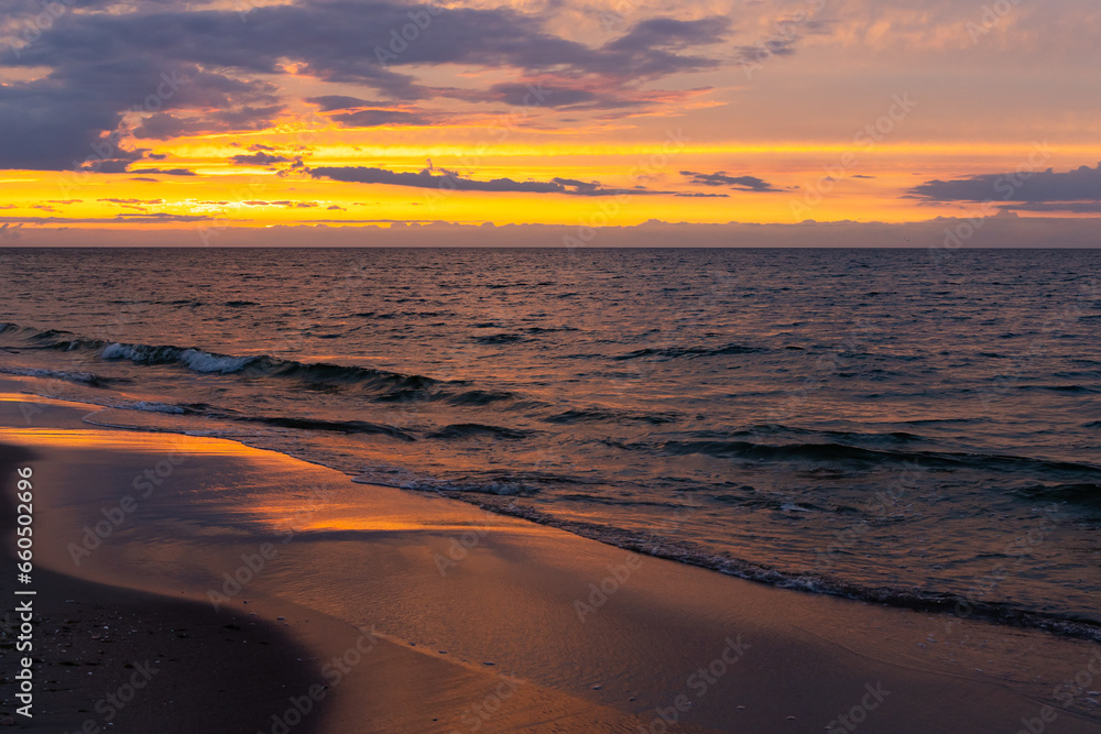Fototapeta premium Orange sunset over the sea. Waves crashing onto the beach, dramatic sky. A static shot taken in the early evening