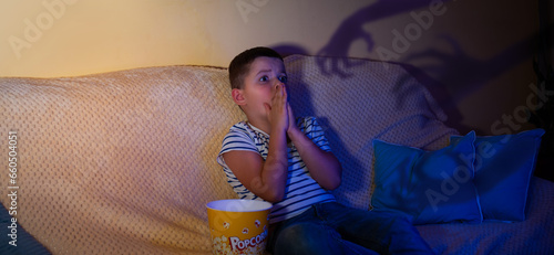 A little boy covered his mouth with his hands in horror and with big eyes watches a movie in a dark room with popcorn.