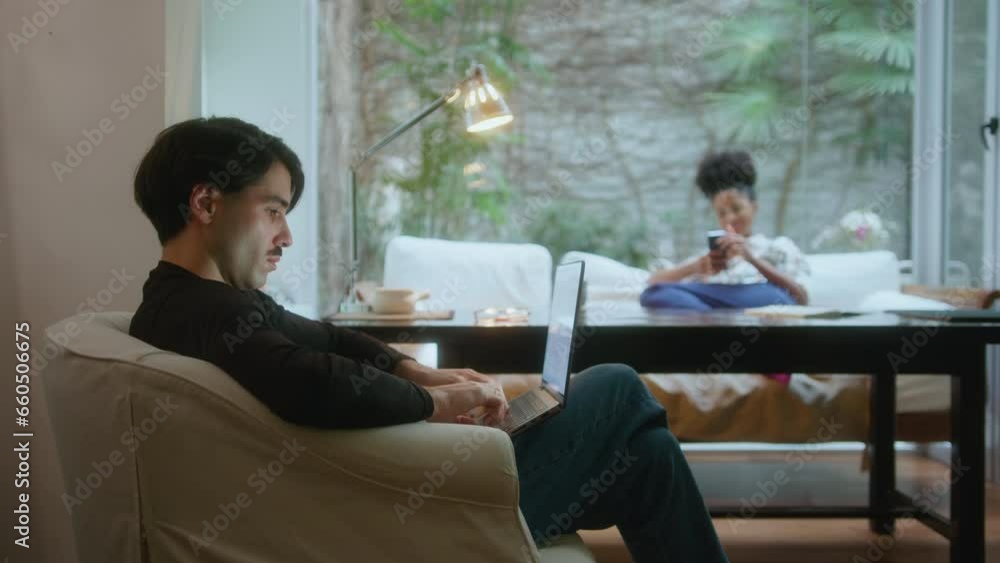 Young man sitting in armchair in living room and working on laptop, wife resting on sofa in the background. Side view, selective focus Young man sitting in armchair in living room and working on laptop, wife resting on sofa in the background. Side view, selective focus