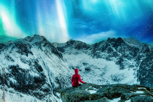 Mountaineer in red jacket sitting on the cliff with Aurora borealis over snowy mountain on Ryten in Lofoten Islands photo