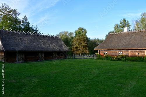 A view of an old wooden hut, shack, or stable surrounded with growing flowers, herbs, a wooden fence, and ancient farming equipment seen next to a well maintained lawn, pastureland, or meadow