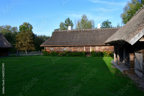 A view of an old wooden hut, shack, or stable surrounded with growing flowers, herbs, a wooden fence, and ancient farming equipment seen next to a well maintained lawn, pastureland, or meadow