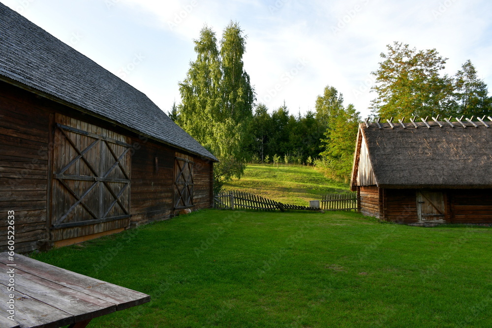 Obraz premium A view of an old wooden hut, shack, or stable surrounded with growing flowers, herbs, a wooden fence, and ancient farming equipment seen next to a well maintained lawn, pastureland, or meadow