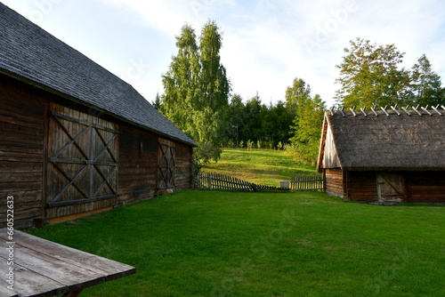 A view of an old wooden hut, shack, or stable surrounded with growing flowers, herbs, a wooden fence, and ancient farming equipment seen next to a well maintained lawn, pastureland, or meadow