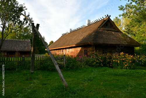 A view of an old wooden hut, shack, or stable surrounded with growing flowers, herbs, a wooden fence, and ancient farming equipment seen next to a well maintained lawn, pastureland, or meadow