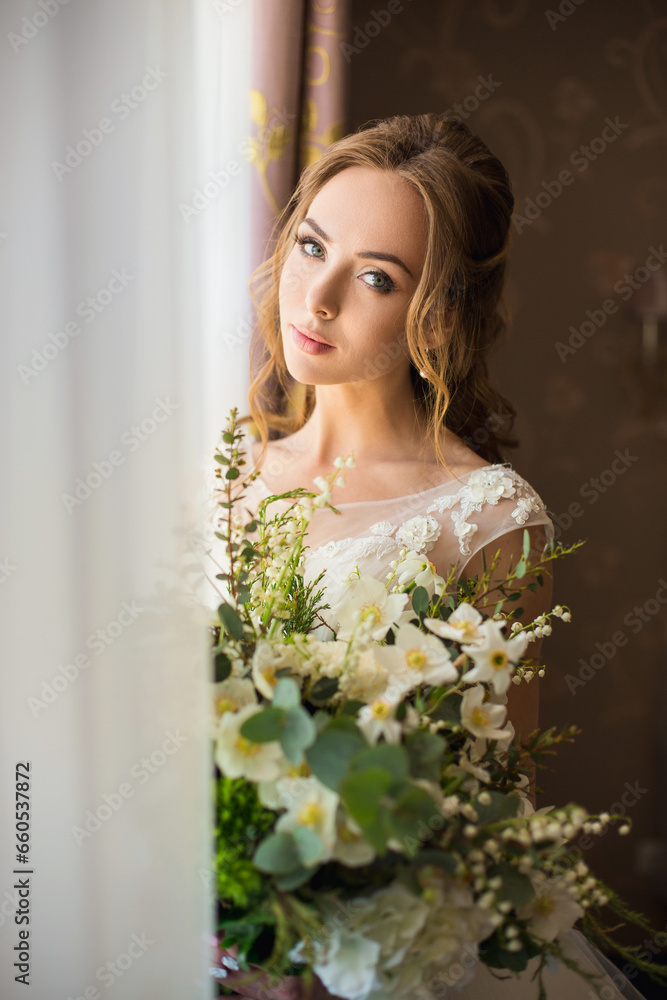 Middle-length portrait of a beautiful young slim sexy caucasian bride in fashionable trendy white gown holding a bouquet of green and white flowers in her hands, standing near window in hotel venue Middle-length portrait of a beautiful young slim sexy caucasian bride in fashionable trendy white gown holding a bouquet of green and white flowers in her hands, standing near window in hotel venue