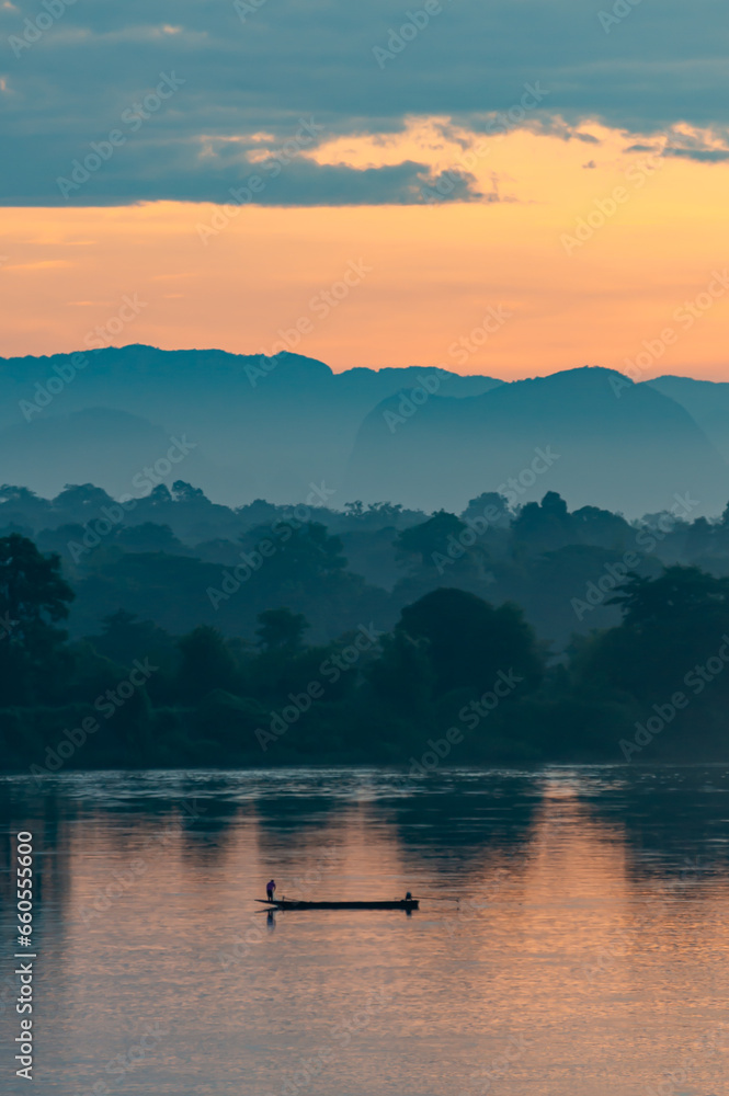 Silhouette of a fishing boat in the Mekong River, bordering Thailand and Laos, in the morning. The background is mountains. There is soft yellow sunlight shining, vertical image suitable for postcards