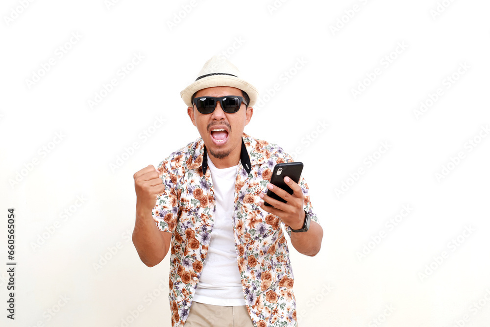 Excited happy adult Asian man tourist holding a cell phone while clenching hand. Concept of travel. Isolated on white Excited happy adult Asian man tourist holding a cell phone while clenching hand. Concept of travel. Isolated on white