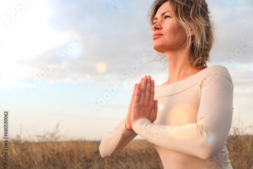 A young woman meditates in nature, with the sky as a backdrop, practicing yoga with her hands in a prayer pose.