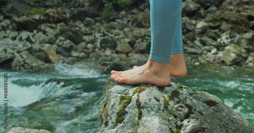 Barefoot woman on rocky shore of river bank. Healthy beautiful female legs close-up. Bare feet girl rests after a hike in the mountains.