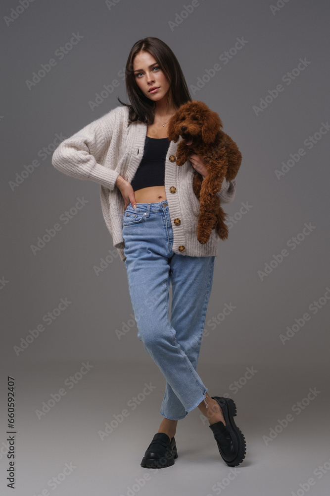 Stylish young brunette woman in casual wear cradling her cherished brown toy poodle, posed before a gray studio backdrop