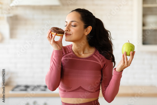 Fitness woman choosing donut instead of healthy apple in kitchen