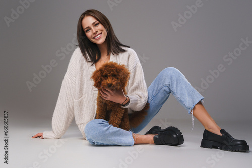 Portrait of a smiling brunette sitting on the floor in casual attire, embracing her cherished toy poodle in a gray studio setting