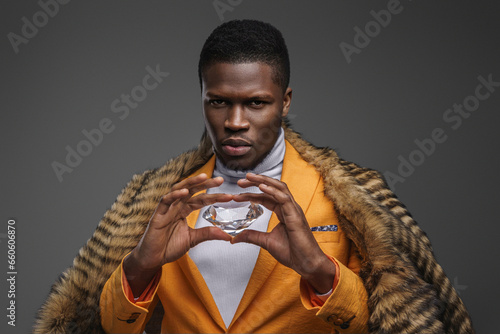 Dapper dark-skinned man adorned in a stunning yellow blazer and draped in a wild animal pelt, showcasing an enormous diamond on a gray background