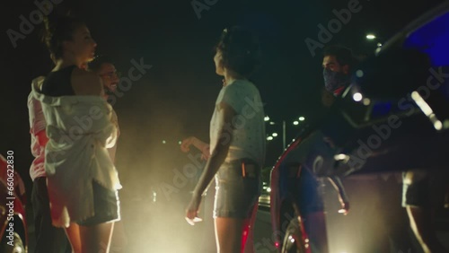 Group of young people dancing near muscle car . Crowd of stylish fans having fan during car races . Young people partying in outdoors at night. Competition of Beautiful american sport cars Slow motion