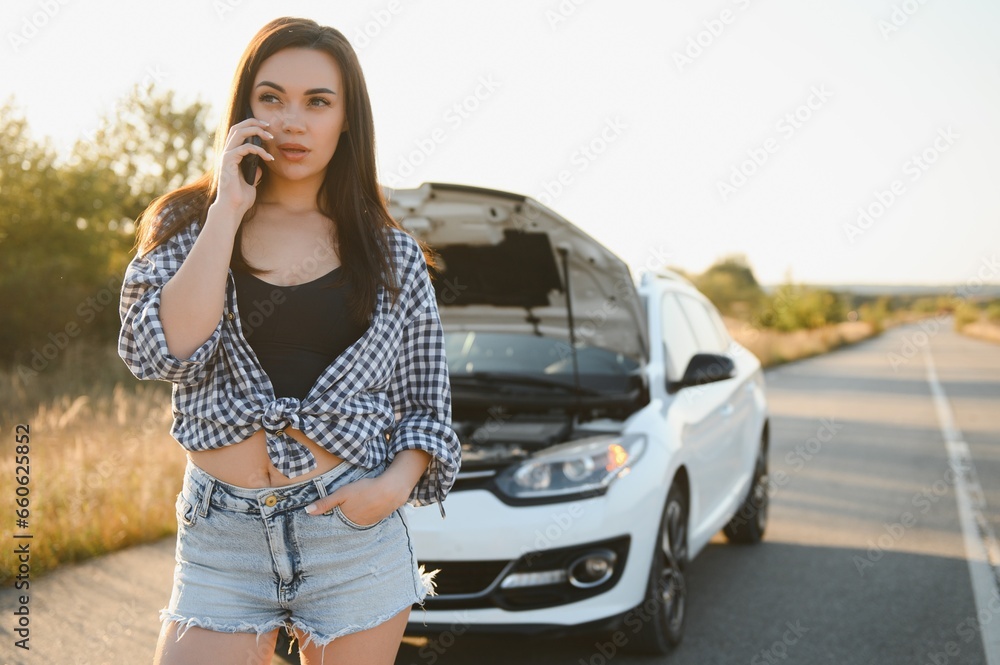The young girl broke the car and she opened the hood and tried to repair the car on the road. A woman calls on a smartphone with a car evacuation service The young girl broke the car and she opened the hood and tried to repair the car on the road. A woman calls on a smartphone with a car evacuation service