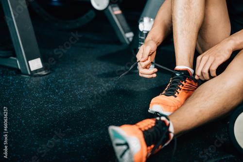 A fit and muscular male is tying his shoelace on the gym floor, surrounded by dumbbells and other workout equipment. The shot emphasizes his commitment to fitness and bodybuilding.