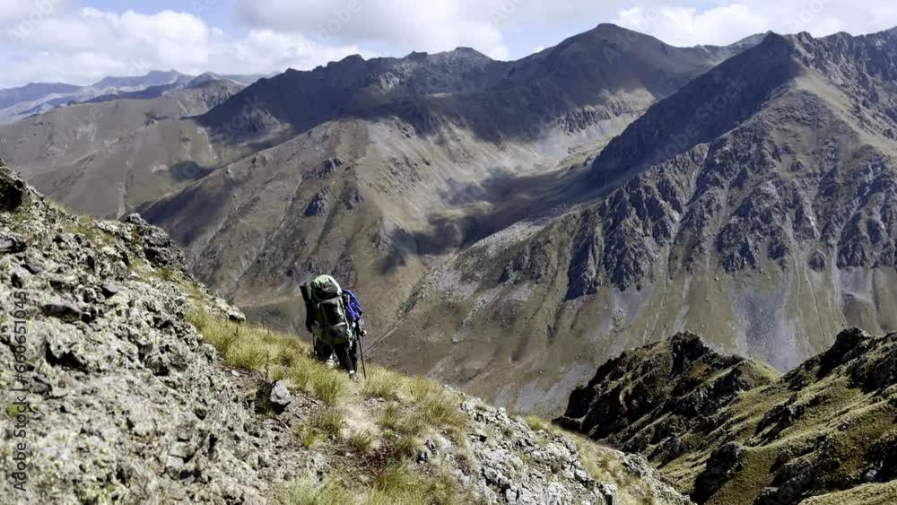 Two hikers are walking through difficult mountainous terrain on a cliff slope, with peaks of rocky mountains visible in the background. Nature and beautiful mountain views. Travelers with backpacks Two hikers are walking through difficult mountainous terrain on a cliff slope, with peaks of rocky mountains visible in the background. Nature and beautiful mountain views. Travelers with backpacks