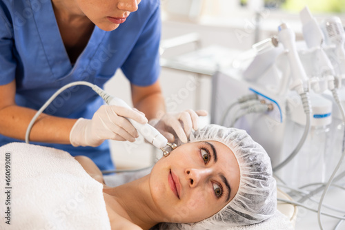 Young woman cosmetologist performs hardware facial microcurrent procedure to young female patient