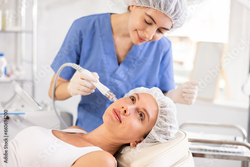 Young female cosmetologist performs cleansing hardware facial procedure on young female patient