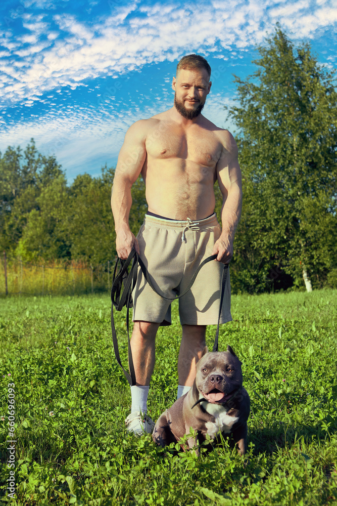 An American Bully walks accompanied by bearded young man in shorts and no shirt.