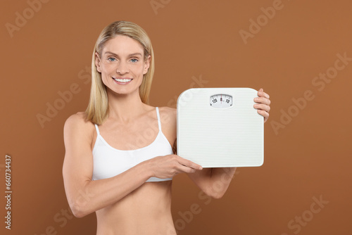 Slim woman holding scales on brown background. Weight loss
